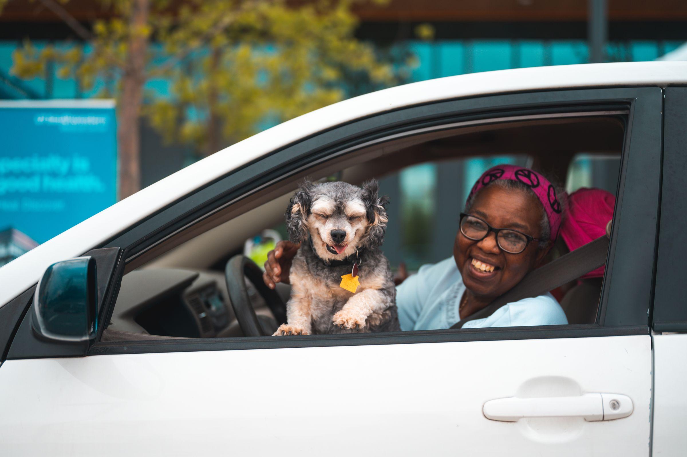 An older lady beaming at the wheel of her car, her dog at the open window