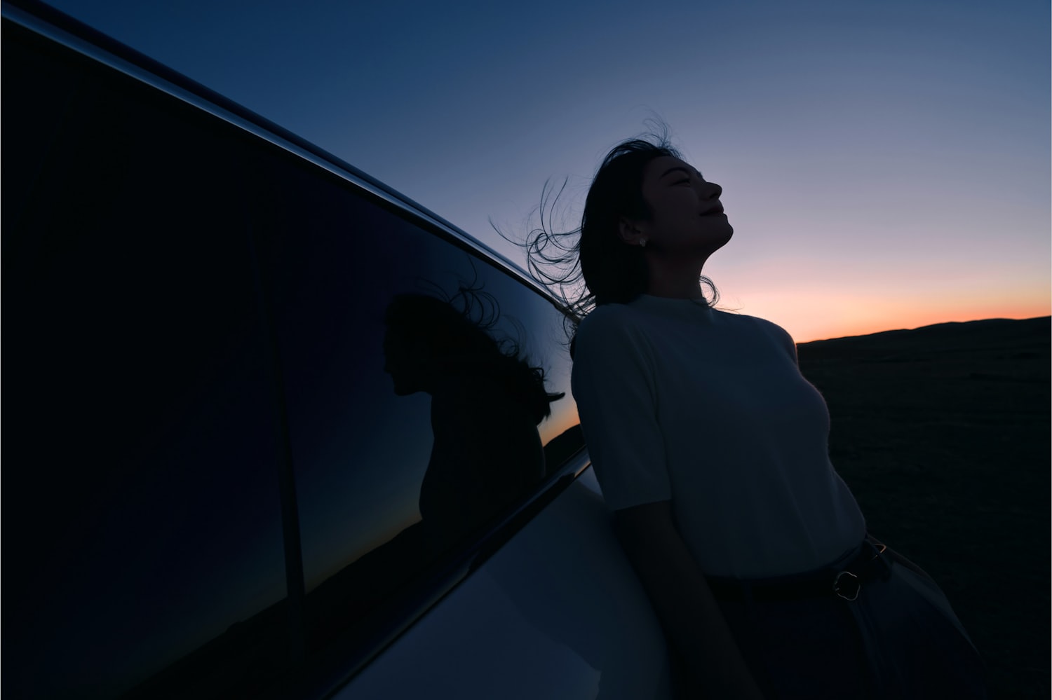 A woman leaning against her car at dusk, calm and at ease
