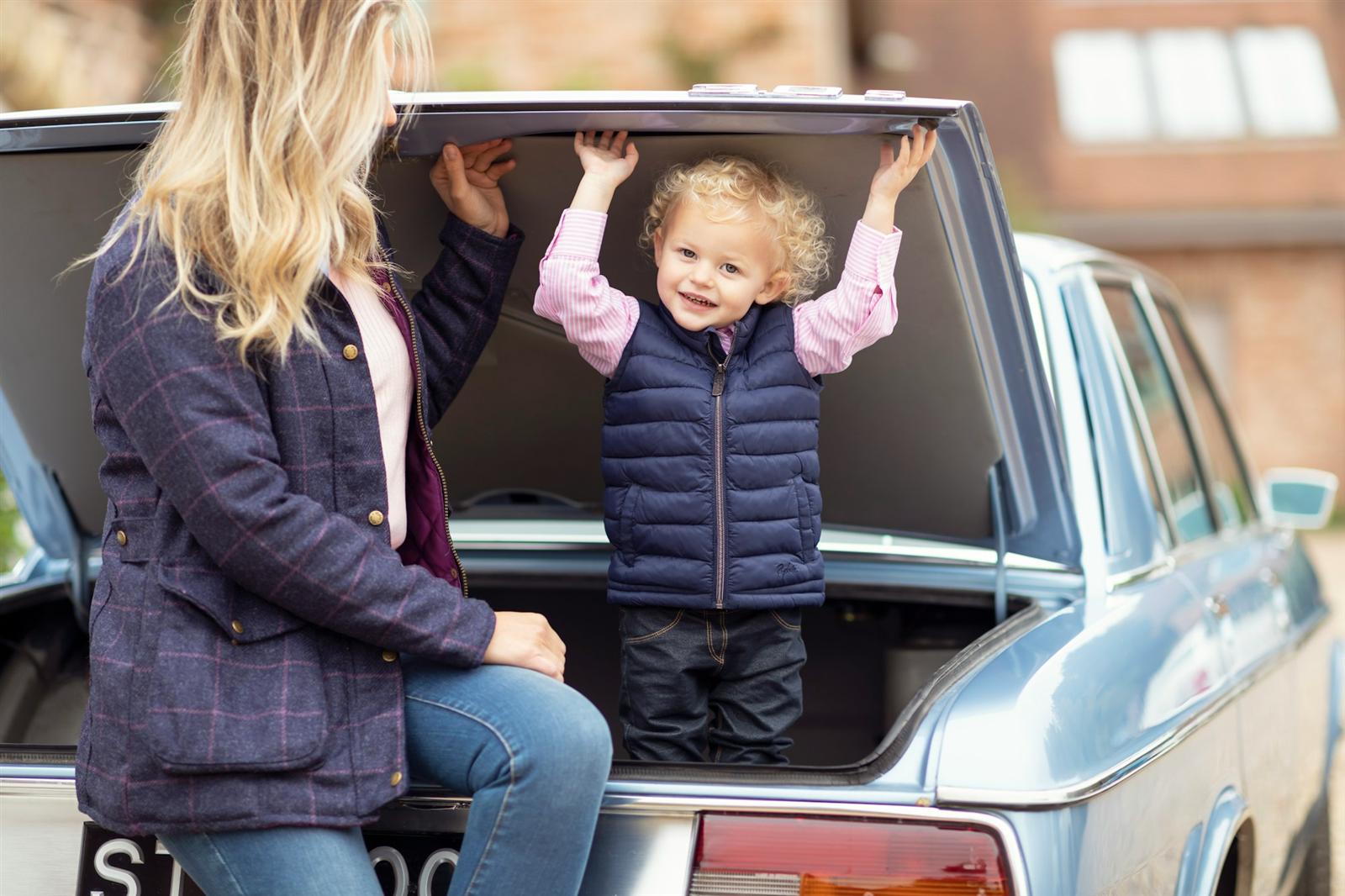 A toddler standing in the open boot of the family car while mum keeps a hand close