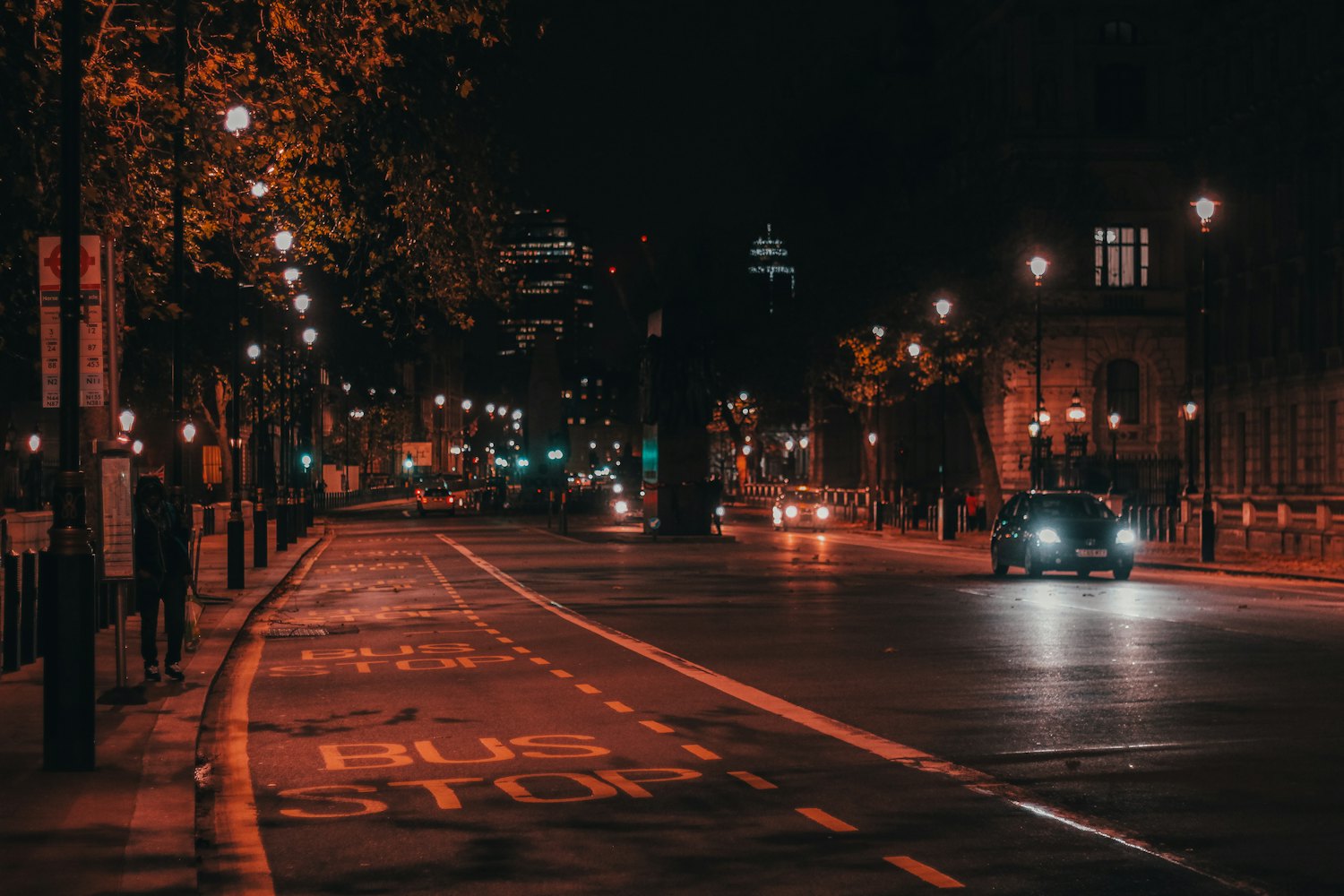 A dark, empty city street at night with a single car driving away under street lamps